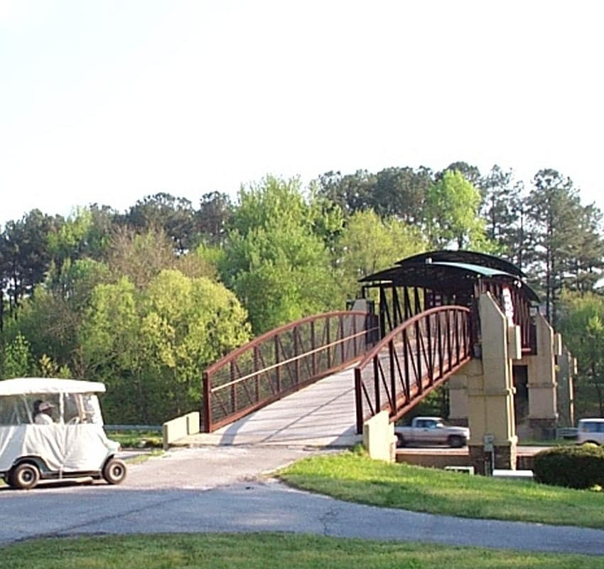 A red metal and concrete pedestrian bridge spans over a small road, surrounded by lush greenery and tall trees. A white golf cart, possibly from the nearby vet clinic, is driving on the road underneath the bridge. The sky is clear, adding to the serene outdoor setting.